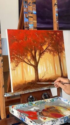 a woman is painting on an easel with red trees in the woods behind her