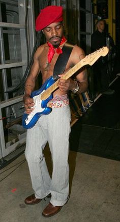 a man standing in front of a building with a guitar on his chest and red bandanna around his neck