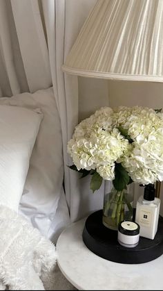 white flowers in a vase on a table next to a lamp and bed with pillows