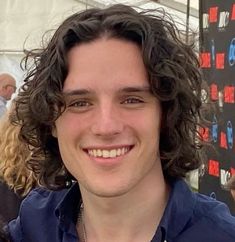 a man with curly hair smiling at the camera while wearing a blue shirt and silver necklace