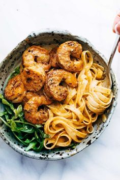 a bowl filled with pasta and shrimp on top of a white marble counter next to a hand holding a fork