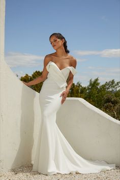 a woman in a white wedding dress leaning against a wall with her hands on her hips