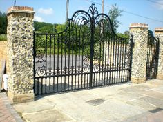 an iron gate with stone pillars on the side of a road in front of a brick building