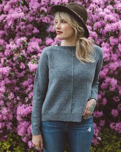 a woman standing in front of purple flowers wearing jeans and a hat with her hands on her hips