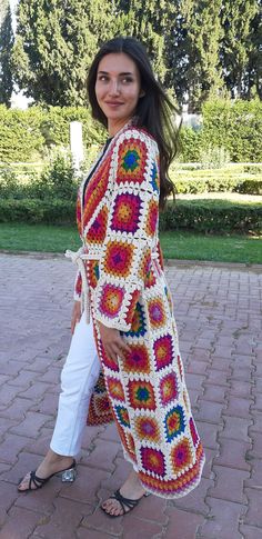 a woman standing on a brick walkway wearing a colorful crocheted cardigan and white pants