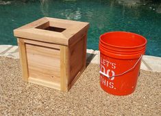 a red trash can next to a wooden box on the ground in front of a swimming pool