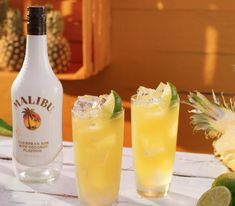 two glasses filled with yellow liquid next to a bottle and pineapple on a table