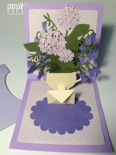 a purple and white greeting card with flowers in a vase on the front, cut out from paper