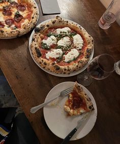 two pizzas sitting on top of white plates next to wine glasses and utensils