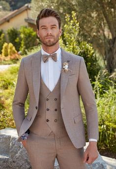 a man wearing a tan suit and bow tie standing in front of some bushes with his hands in his pockets