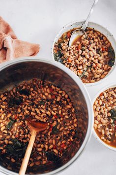 three bowls filled with beans and greens next to a wooden spoon on a white surface