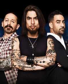 three men with tattoos and piercings standing in front of a dark background, one man has his arms crossed