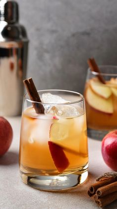 two glasses filled with apple cider next to cinnamon sticks and apples on the table