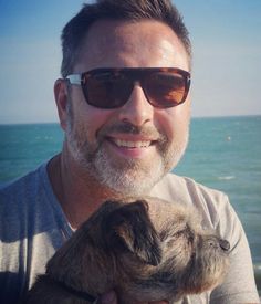 a man in sunglasses holding a small dog by the water's edge on a sunny day