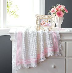 a crocheted baby blanket sitting on top of a dresser next to a vase with flowers