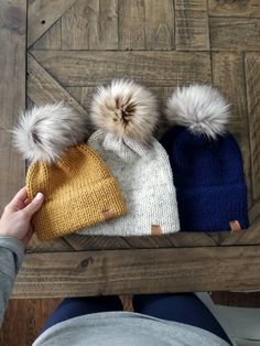 three hats with two pom poms sitting on top of a wooden table next to a person's legs