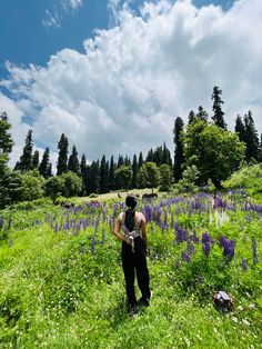 a woman standing in the middle of a lush green field with purple flowers on it