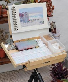 an artist's easel with watercolors and paint on it in front of some potted plants