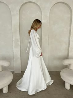 a woman in a white dress standing next to two chairs and a wall with arches