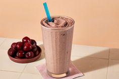 a smoothie in a tall glass next to a bowl of cherries