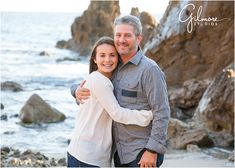 a man and woman hugging each other on the beach near the ocean with rocks in the background