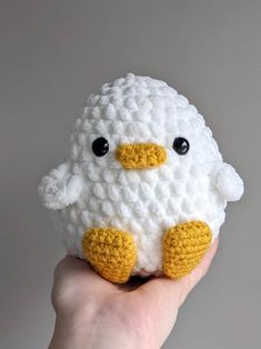 a hand holding a small white crocheted stuffed animal with black eyes and a yellow beak