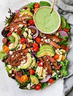 a salad with steak, tomatoes, avocado, corn and dressing on it