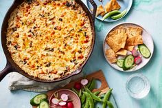 a casserole with cheese and vegetables next to crackers, cucumbers and other foods