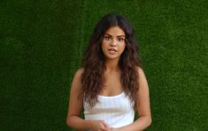 a woman standing in front of a green grass wall with her hands together and looking at the camera