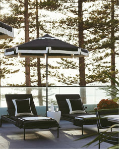 Stylish black lounge chairs with crisp white pillows under a classic black and white striped umbrella, creating a sophisticated poolside look. Perfect for relaxation with a view of the sea and towering pine trees. Market Umbrella, Vintage Look, Vintage Looks, Umbrella