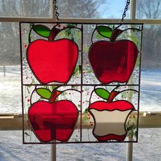 three stained glass apples hanging on a window sill in the snow with trees behind them