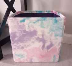 a tie dyed storage bin sits on the floor next to a chair and wall with a black frame