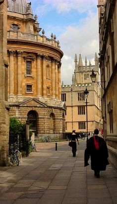 two people walking down the street in front of some buildings with tall spires on them