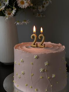 a pink cake with two candles on it and daisies in the vase next to it