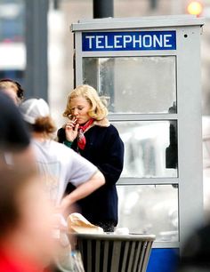 a woman standing in front of a telephone booth talking on her cell phone while people walk by