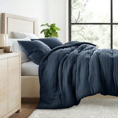 a bed with blue sheets and pillows in front of a large window on a white rug