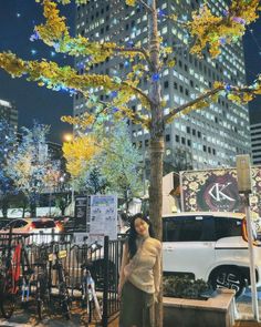 a woman standing next to a tree in front of a tall building at night time