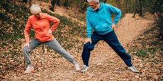 an older man and woman doing stretching exercises on a path in the woods with fallen leaves