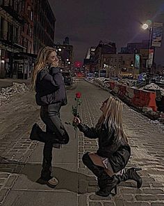 a woman kneeling down next to another woman holding a rose in her hand on the sidewalk