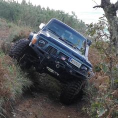 a blue jeep driving down a dirt road next to a tree and grass covered hillside