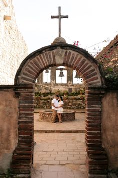 two people sitting on a bench in front of an arch with a cross above them