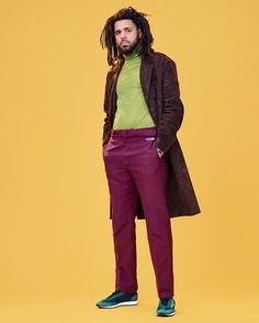 a man with dreadlocks standing in front of an orange background wearing a green shirt and maroon pants