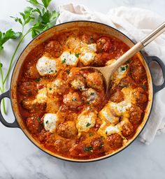 a pot filled with meatballs and cheese on top of a white table cloth next to a wooden spoon