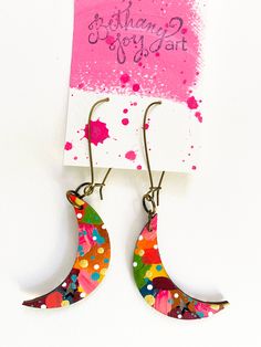 a pair of earrings with colorful designs on them and a pink card in the background