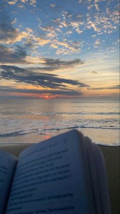 an open book sitting on top of a sandy beach next to the ocean at sunset