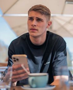 a young man sitting at a table looking at his cell phone and holding a cup