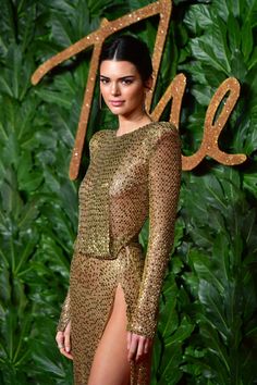 a woman in a gold dress standing on a red carpet with green leaves behind her