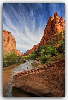 a river flowing through a lush green forest covered hillside next to tall red rock formations