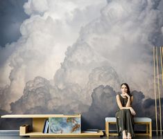 a woman sitting on a bench in front of a wall with clouds painted on it