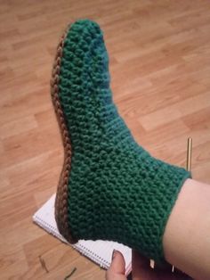 someone wearing green knitted slippers on top of a wooden floor next to a pair of knitting needles